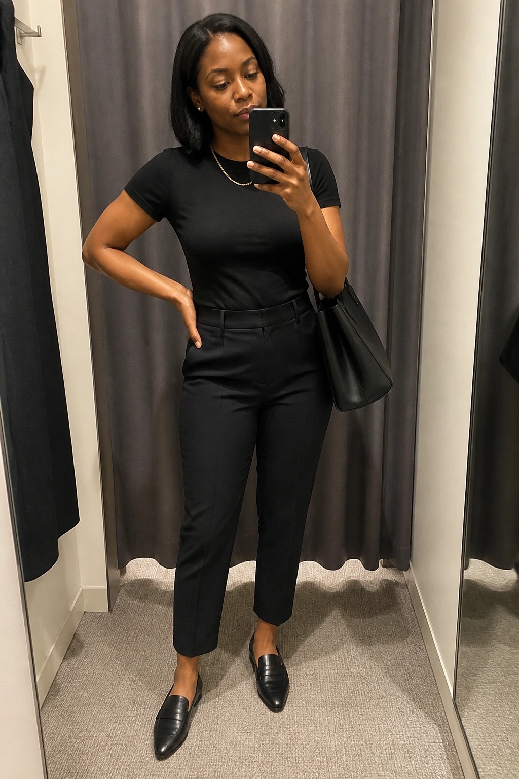 Woman in black top and pants taking mirror selfie while holding handbag in fitting room