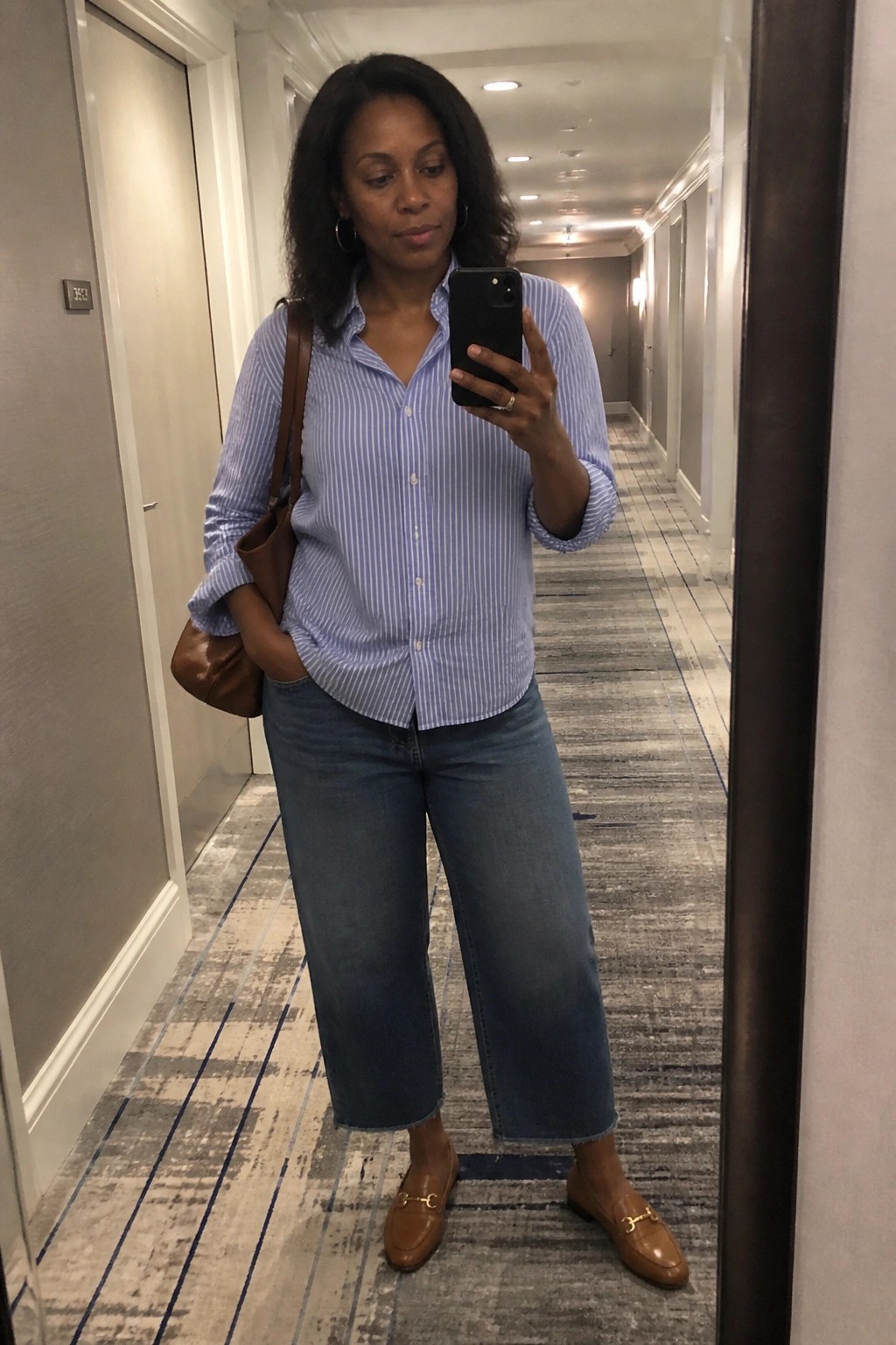 A Black woman wearing a blue and white striped button down shirt tucked into wide leg cropped blue jeans with brown loafers and a brown leather tote bag taking a mirror selfie.
