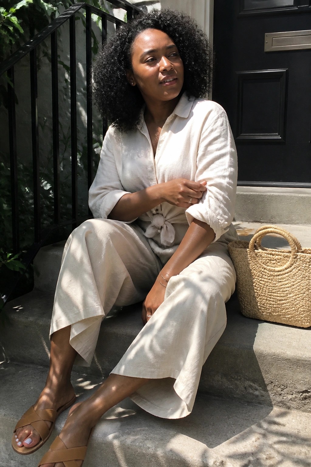 Curly-haired woman in white linen outfit seated on steps beside woven bag