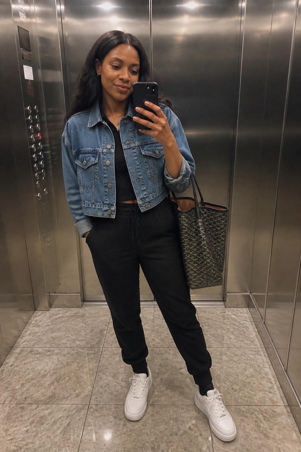 Woman in an elevator wearing a cropped blue denim jacket over a black top with black jogger pants, white sneakers, and a patterned tote bag while taking a mirror selfie.