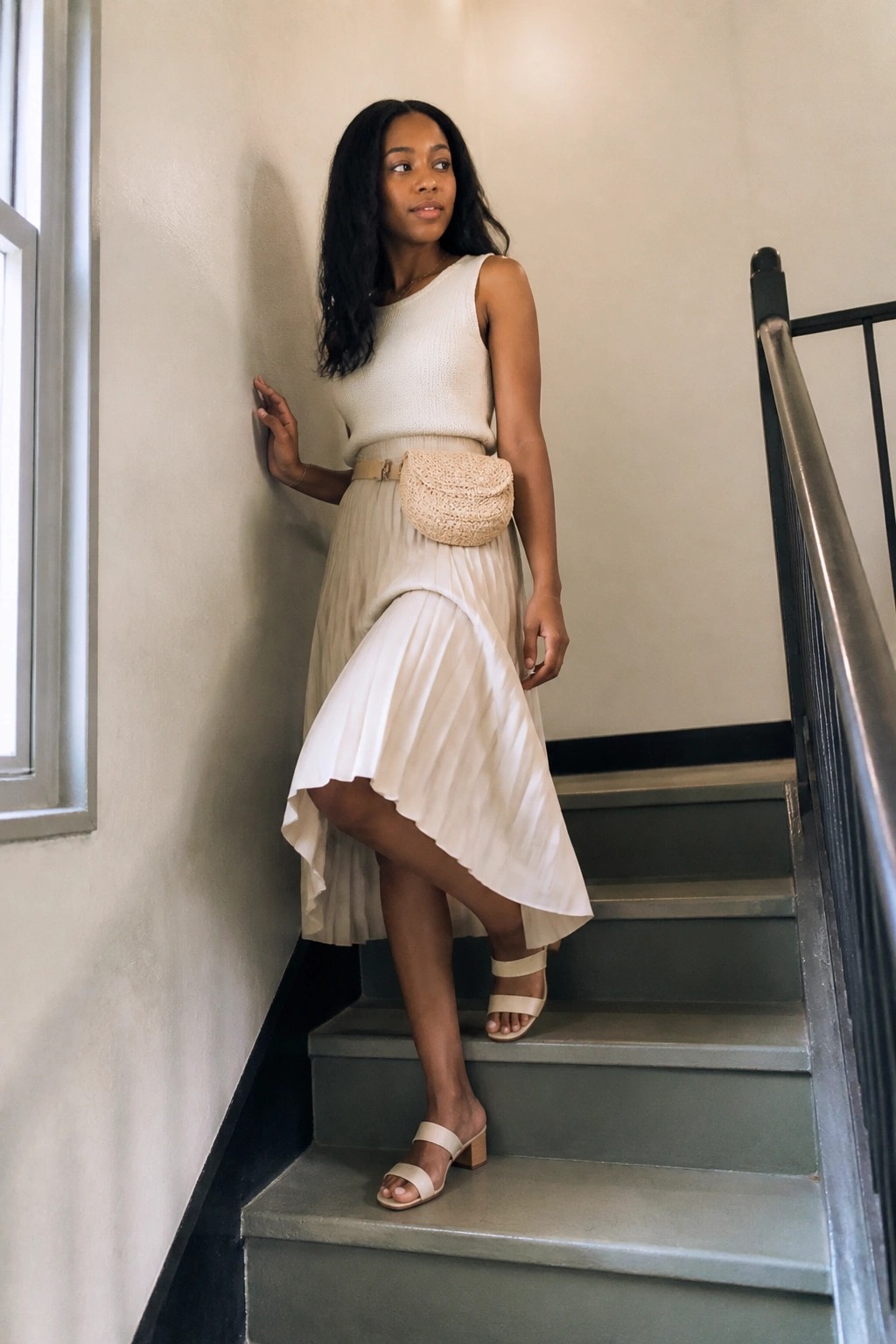 Woman in white top and beige pleated skirt with woven bag on staircase