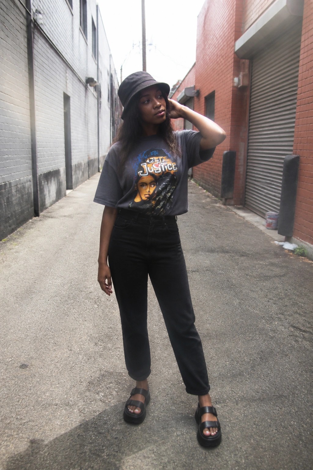 Woman in bucket hat, graphic tee, black jeans, and platform sandals in alley