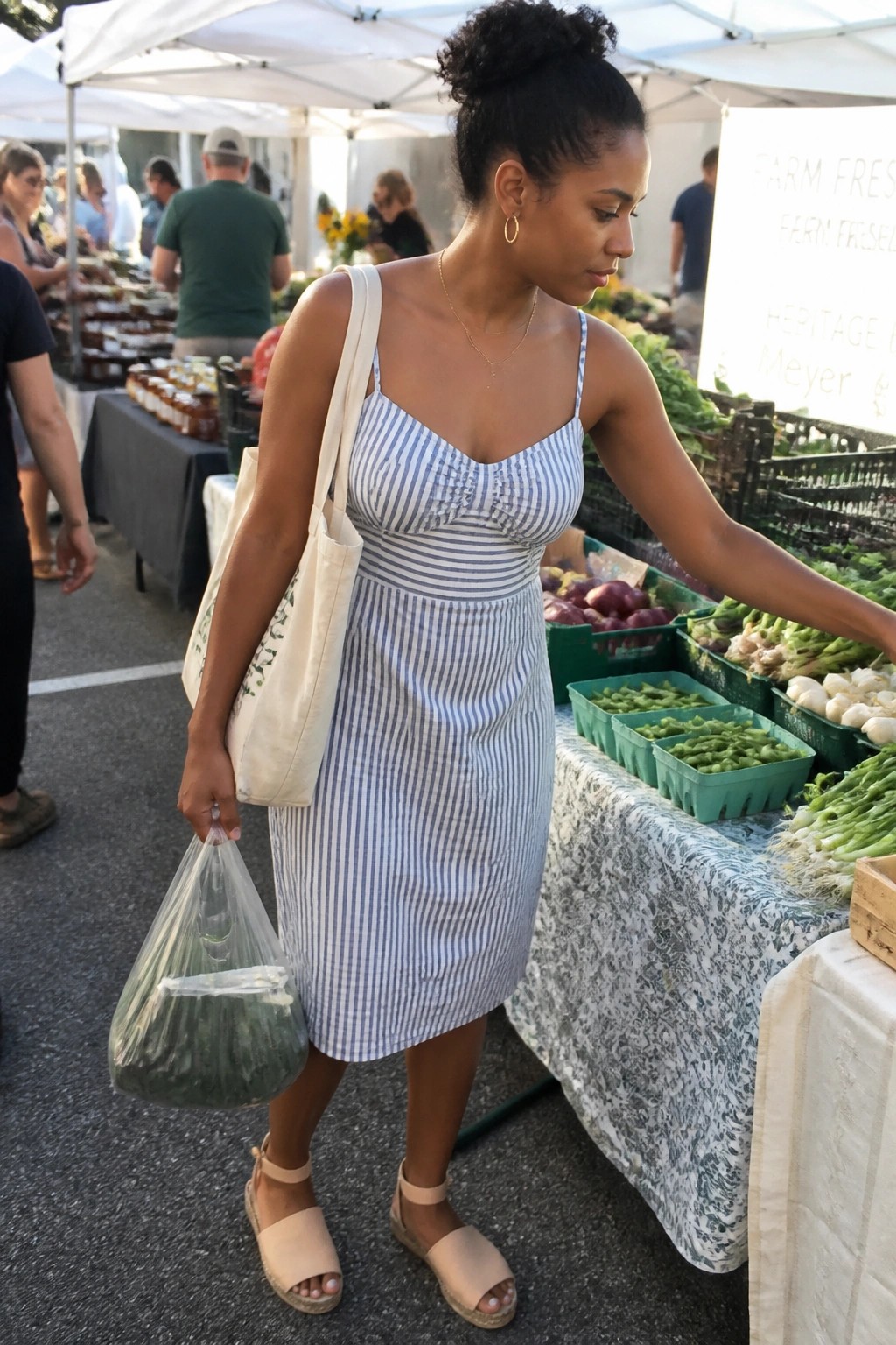 A Black woman wearing a blue and white striped midi dress with thin straps, carrying a canvas tote bag and a plastic bag of greens, paired with beige espadrille sandals at a market stall.