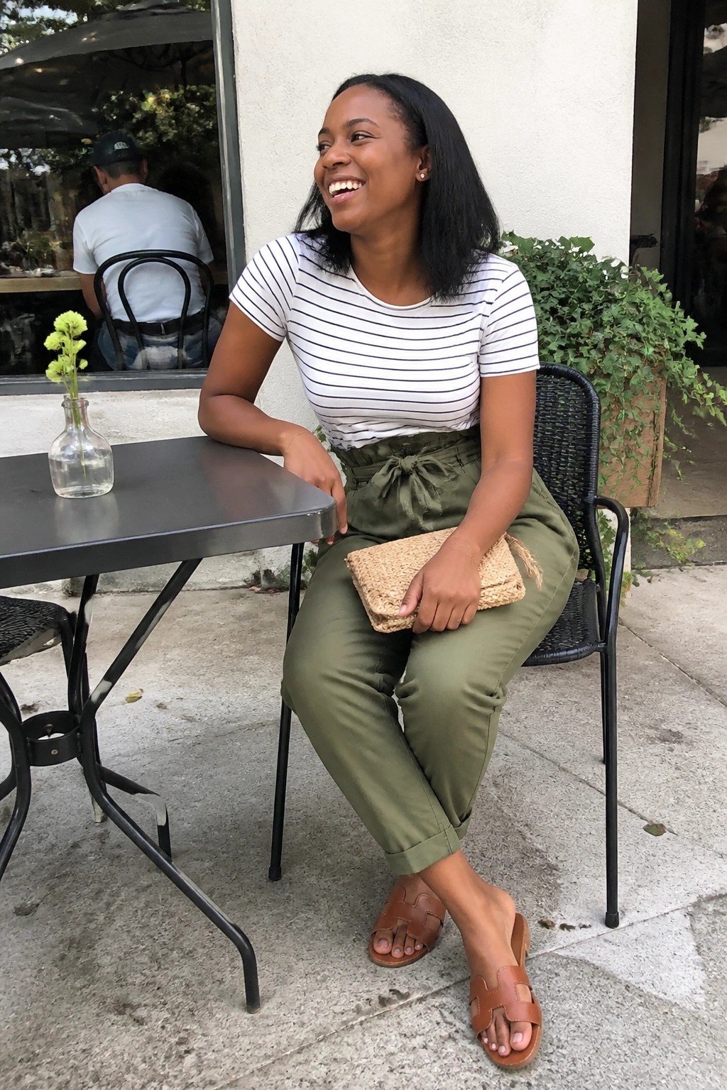 Smiling woman in striped top and green pants holding woven clutch at cafe table