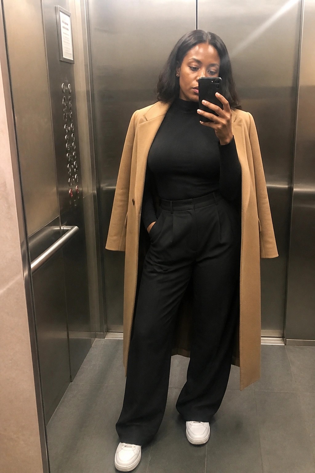 Woman in black turtleneck, tan coat, and wide pants taking elevator selfie.