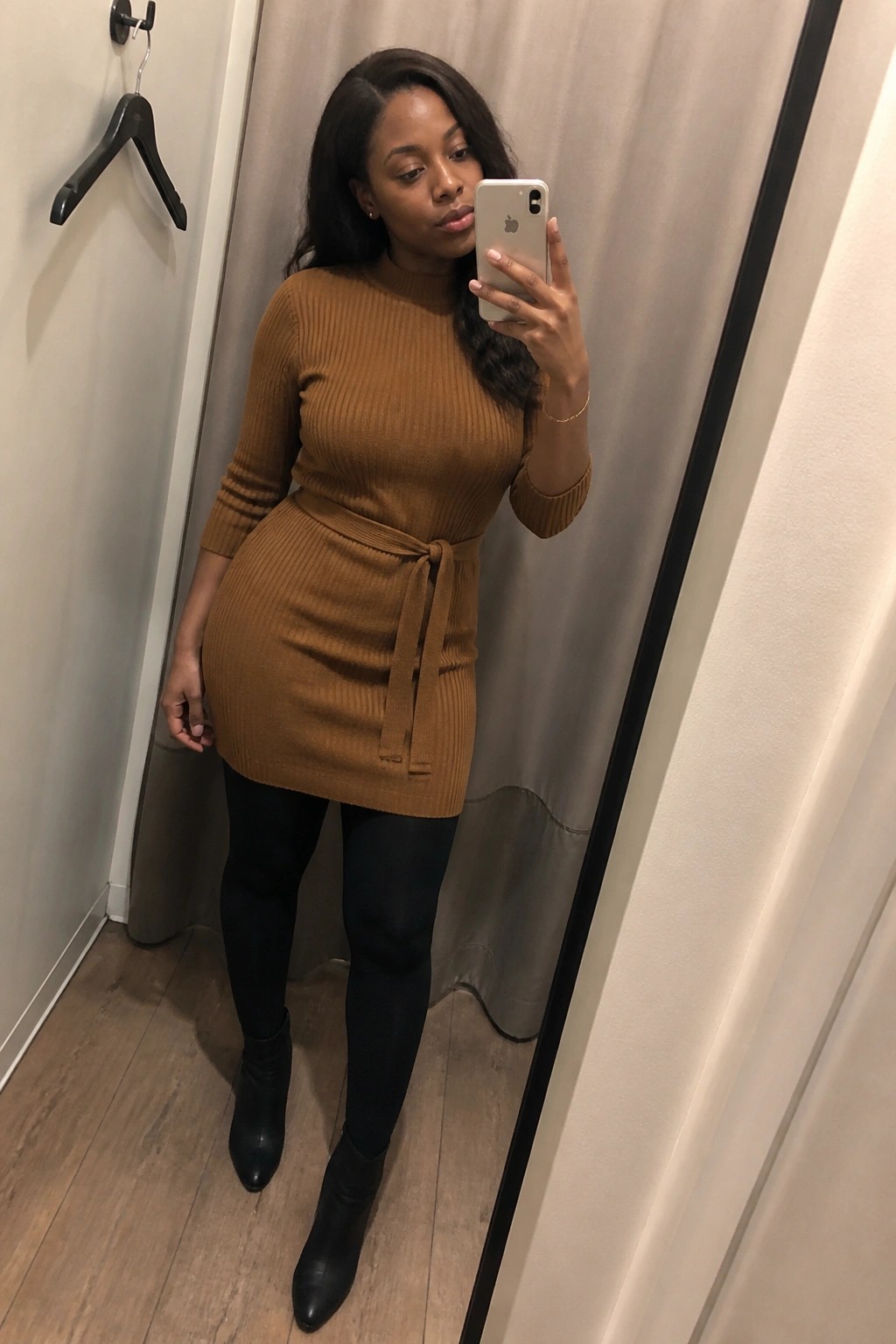 Woman in brown ribbed sweater dress with belt, black boots, holding iPhone mirror selfie