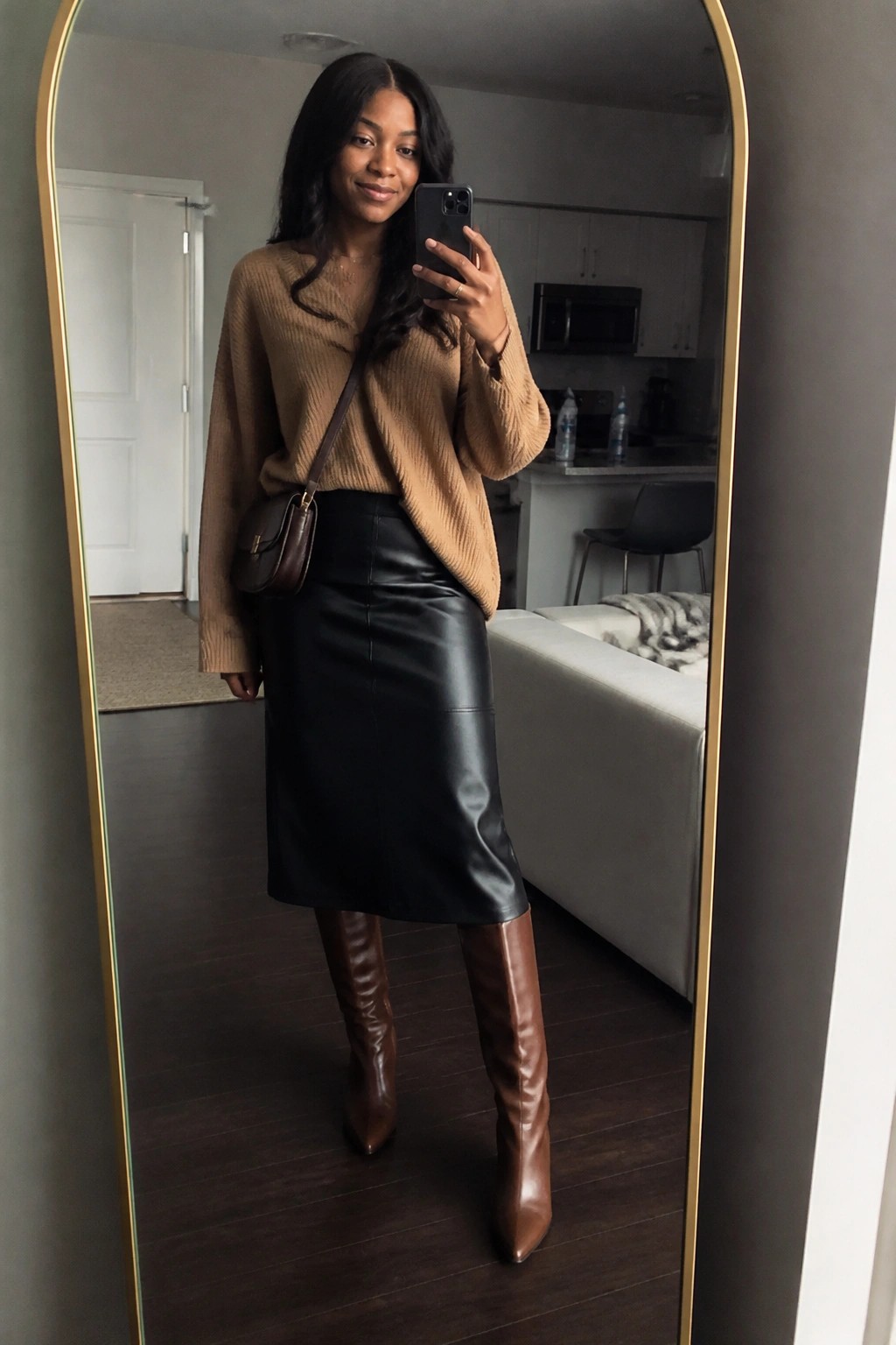 Woman in tan sweater and black leather skirt taking mirror selfie in apartment.