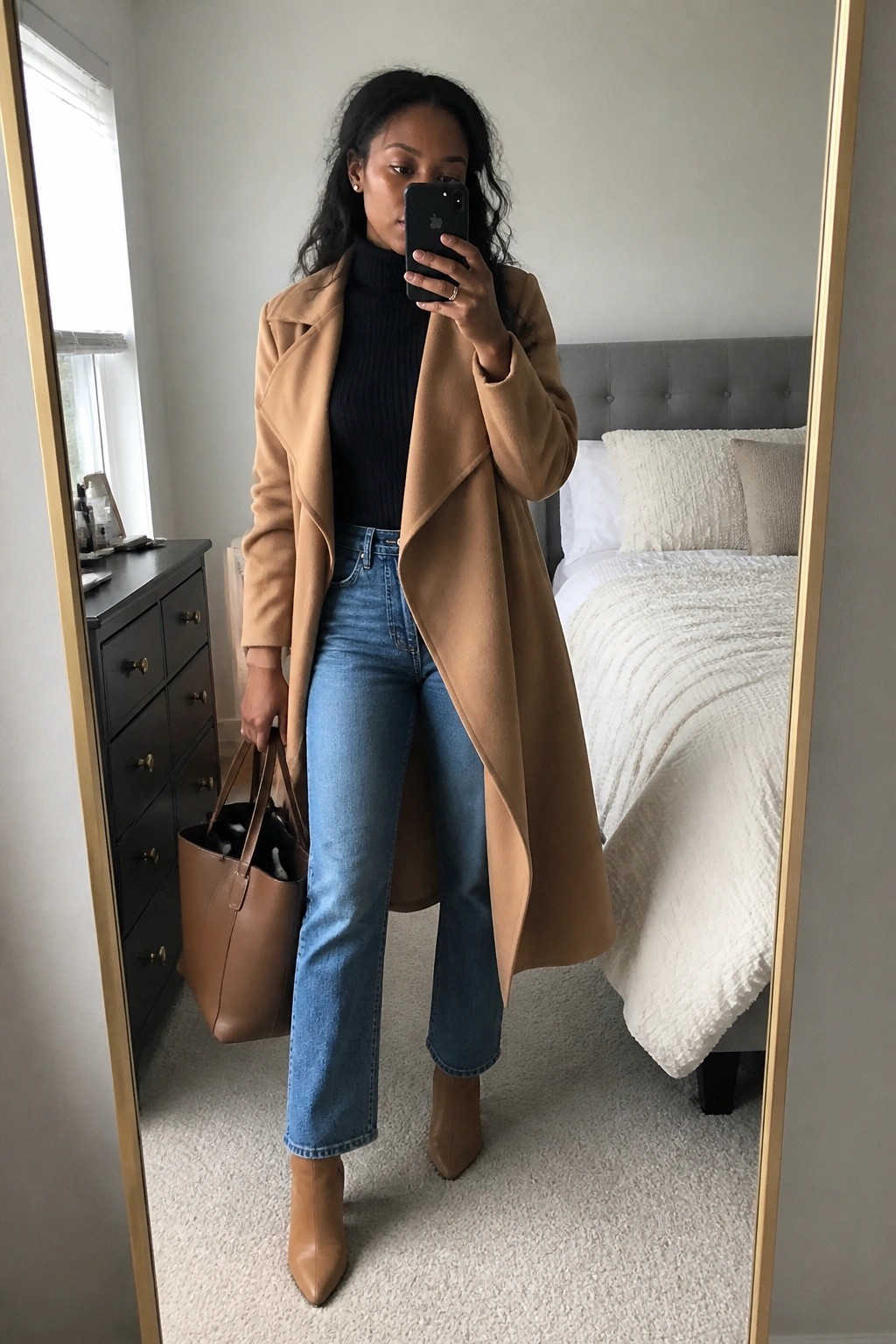 Woman in camel coat and jeans taking mirror selfie while holding brown tote bag