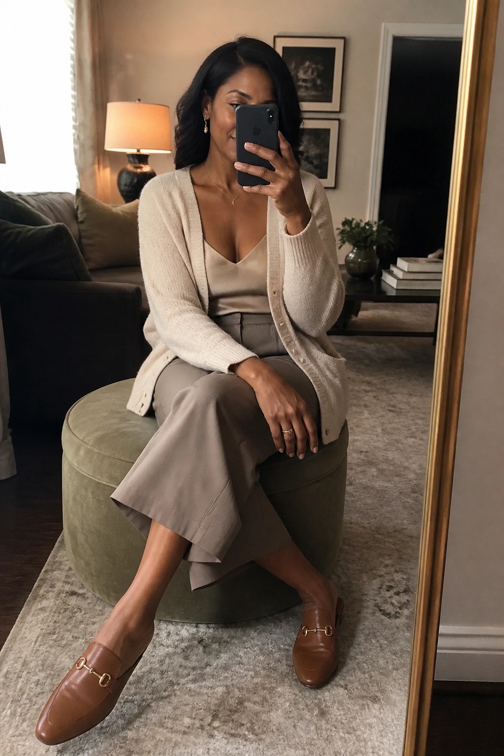 Woman in cream cardigan and taupe pants taking mirror selfie on green ottoman