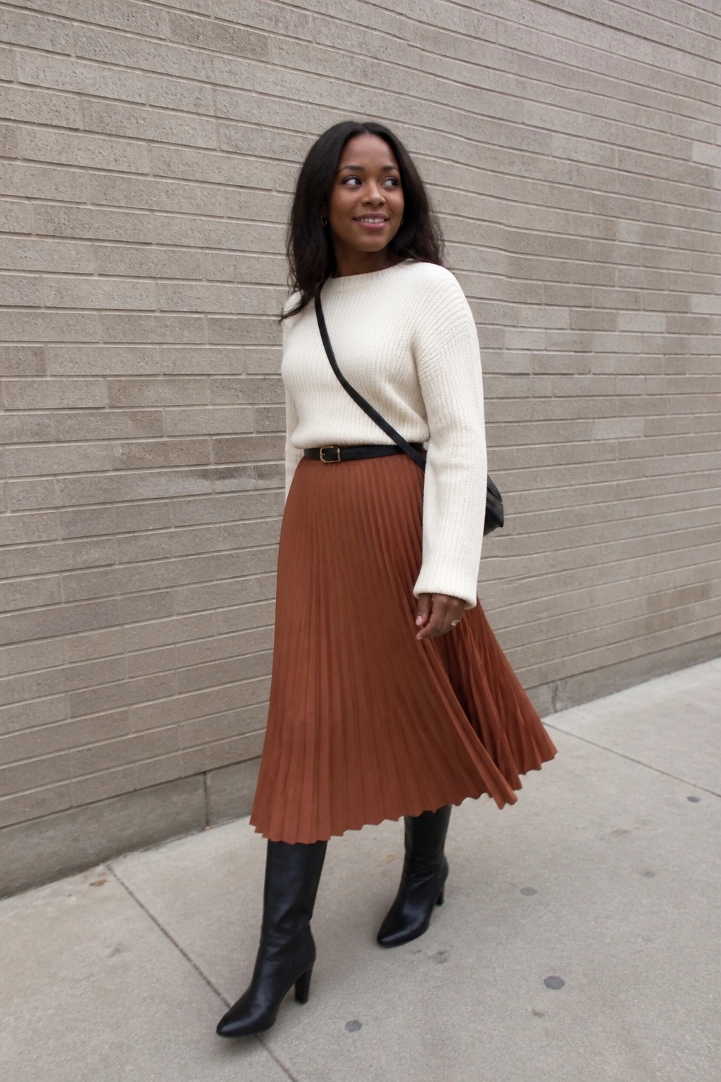 Woman in cream sweater and brown pleated skirt with black boots by brick wall