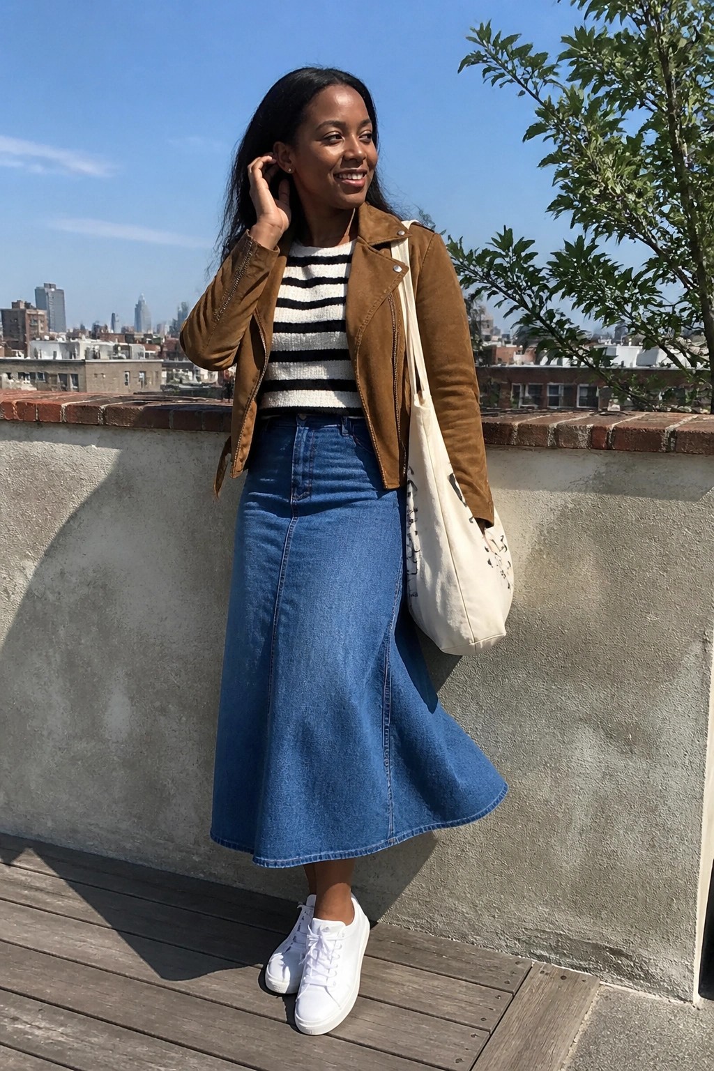 Black woman in brown suede jacket over black and white striped knit top with blue denim midi skirt white sneakers and canvas tote bag standing on rooftop.