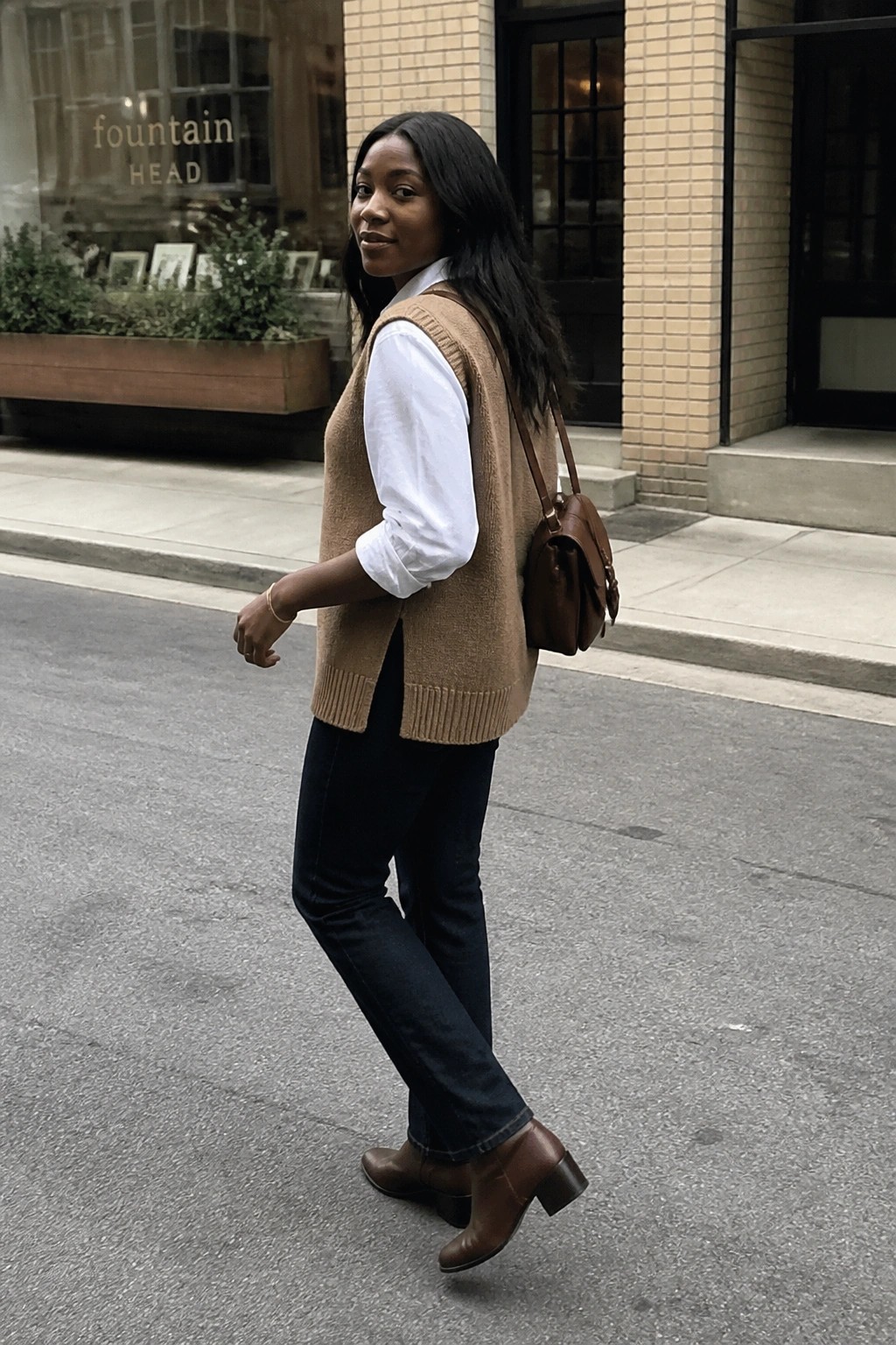 Woman in brown knit vest over white shirt with leather bag on city sidewalk