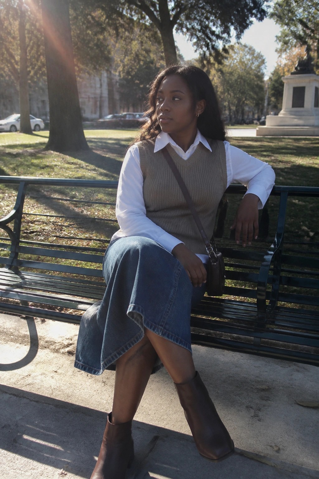 Black woman in beige sweater vest, white shirt, and denim skirt on park bench
