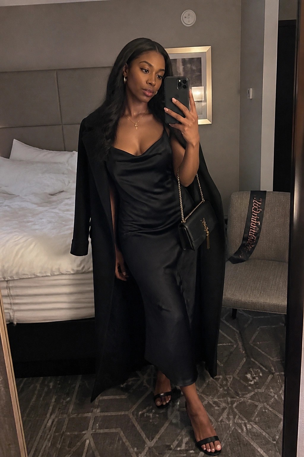 Woman wearing a black satin cowl neck midi slip dress with a long black coat draped over her shoulders, holding a black phone and a small black YSL chain strap bag, paired with black heeled sandals and gold jewelry in a hotel room setting.