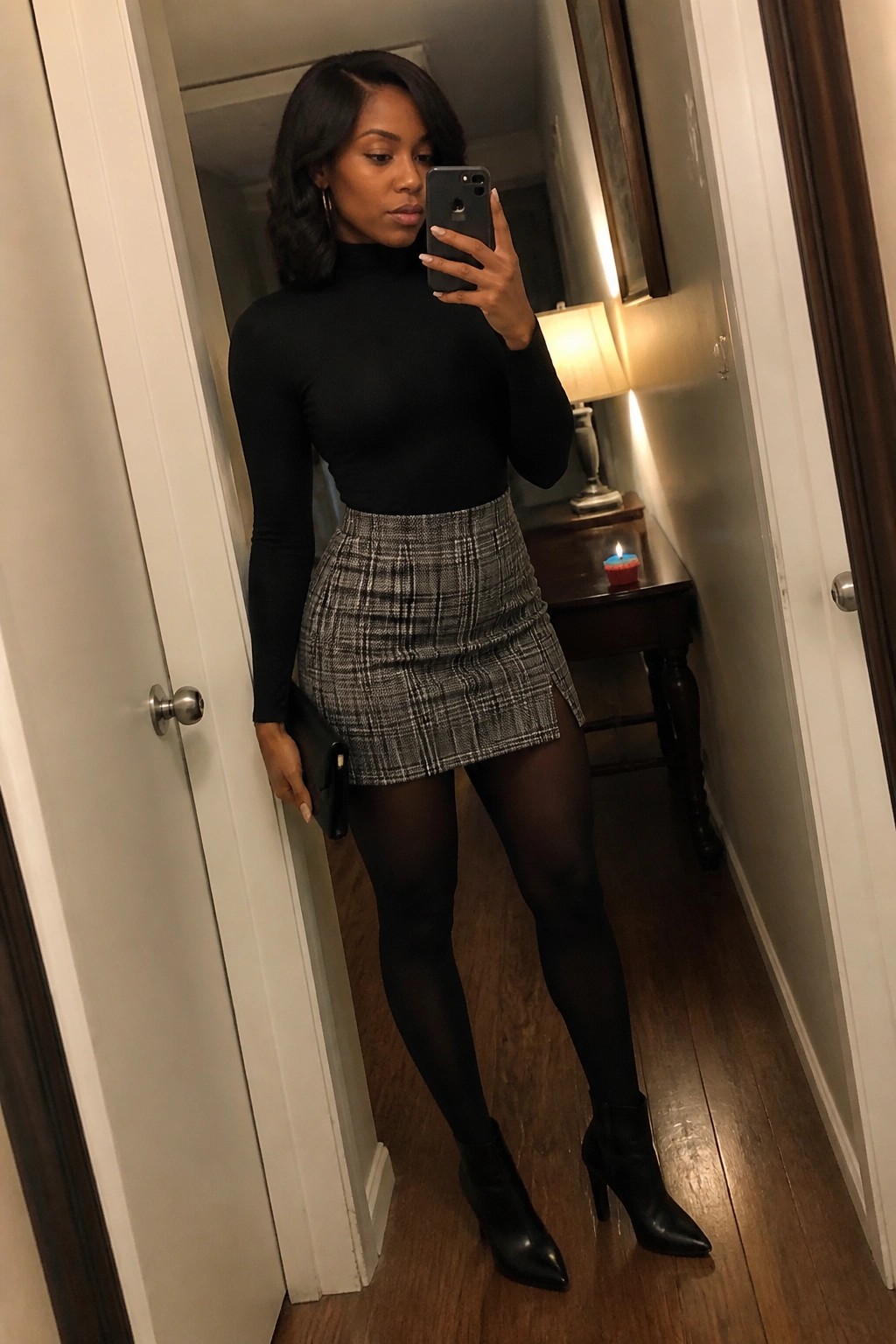 Woman wearing a fitted black turtleneck top, gray black plaid mini skirt with side slit, black tights, and pointed black heeled ankle boots while holding a black clutch and taking a mirror selfie.
