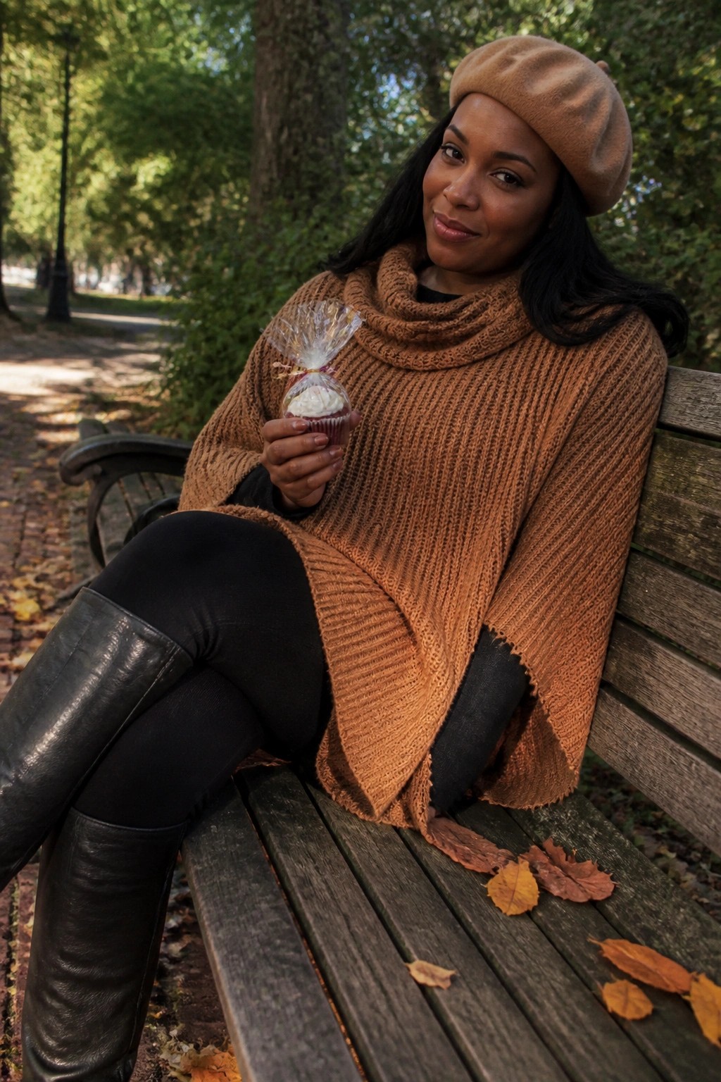 Woman in brown beret and knit poncho holding cupcake on autumn park bench