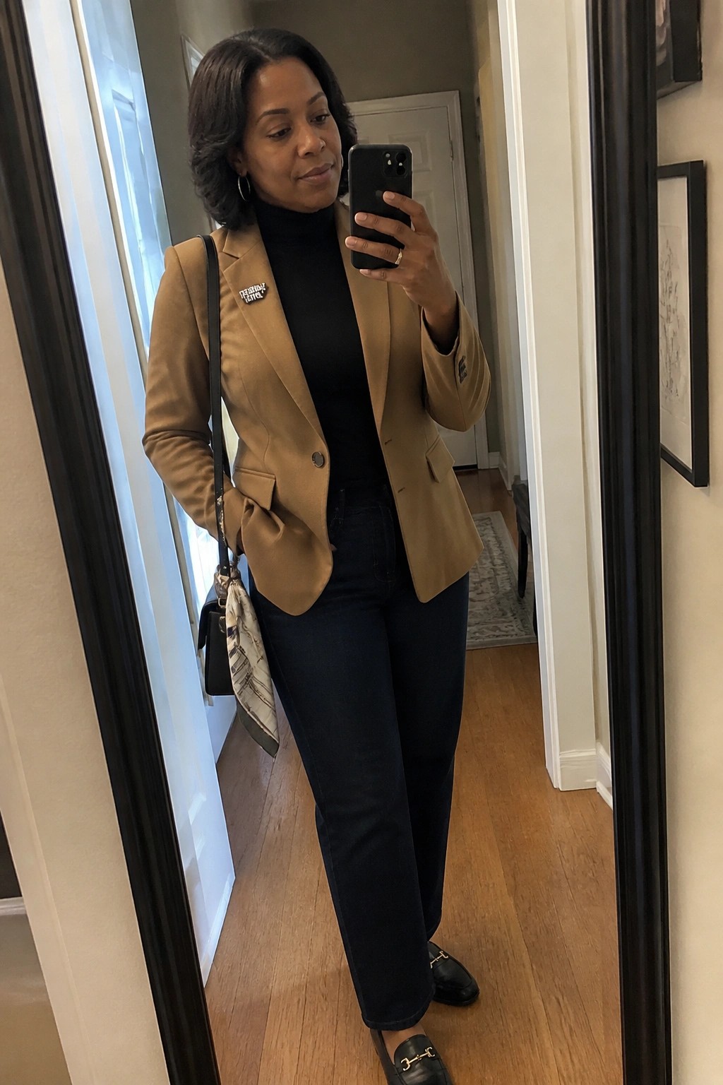 Woman in tan blazer over black turtleneck with jeans and handbag in hallway