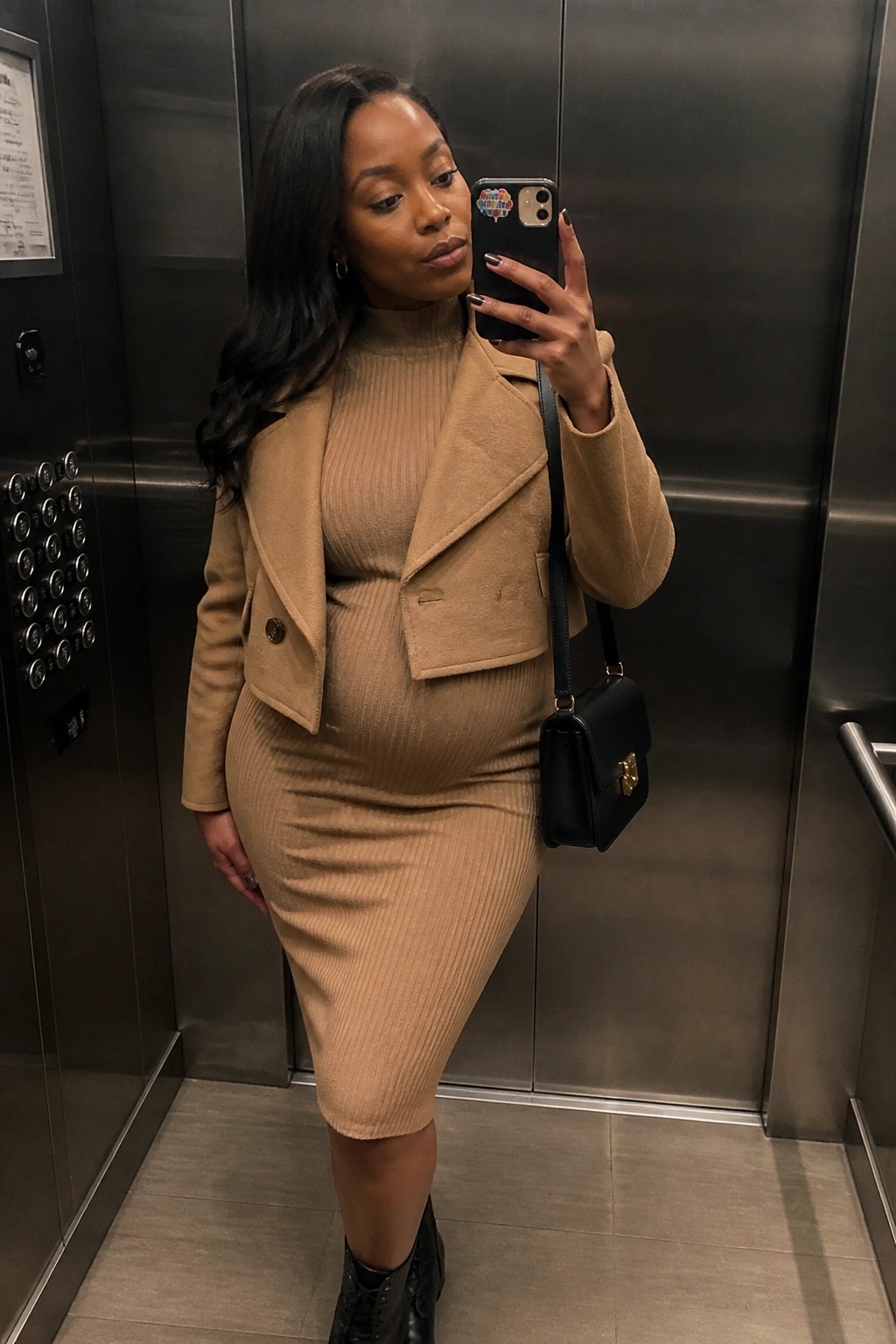 Woman in tan ribbed dress and cropped jacket taking elevator selfie with black bag