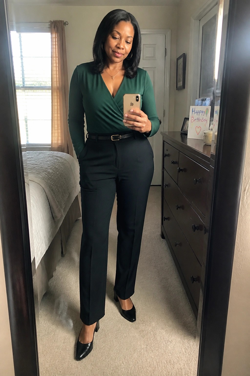 A woman in a dark green long sleeve wrap top tucked into black straight leg trousers with a thin black belt and pointed black heels stands in a bedroom mirror selfie holding a phone.