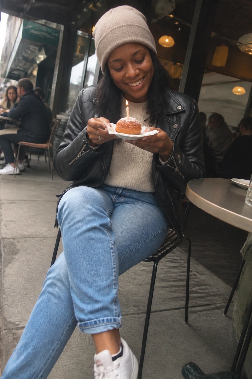 Woman in beanie and leather jacket smiles at donut with lit candle.