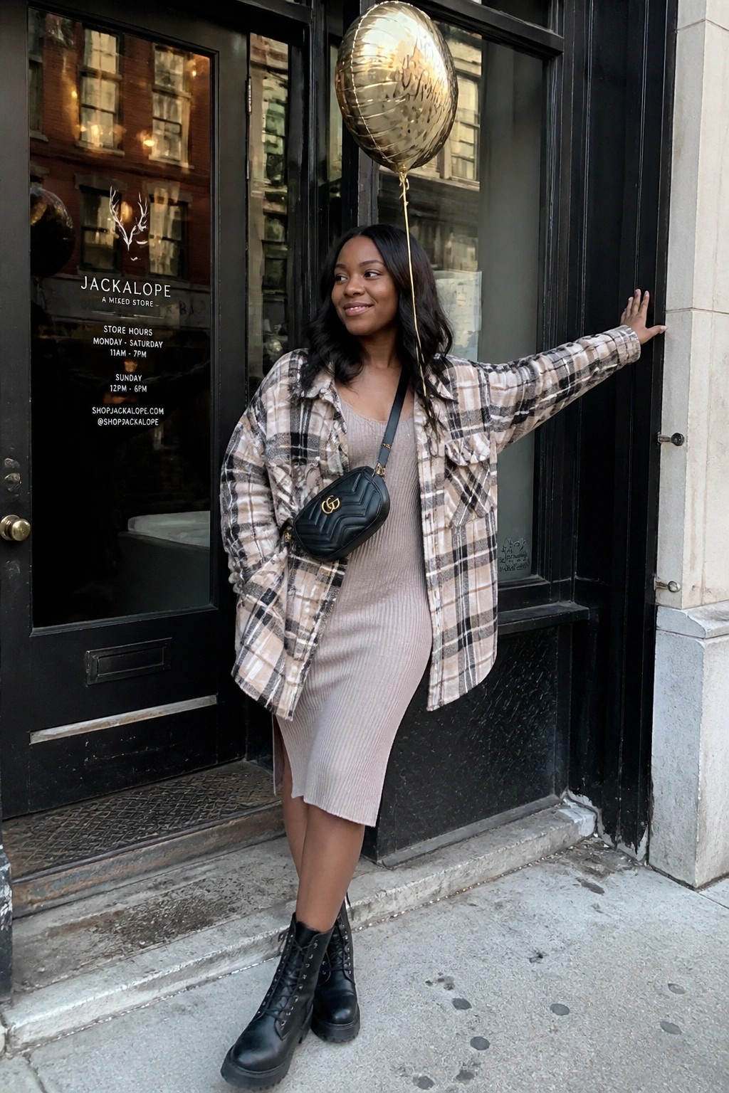 Woman in plaid shirt over beige dress holding gold balloon outside storefront.
