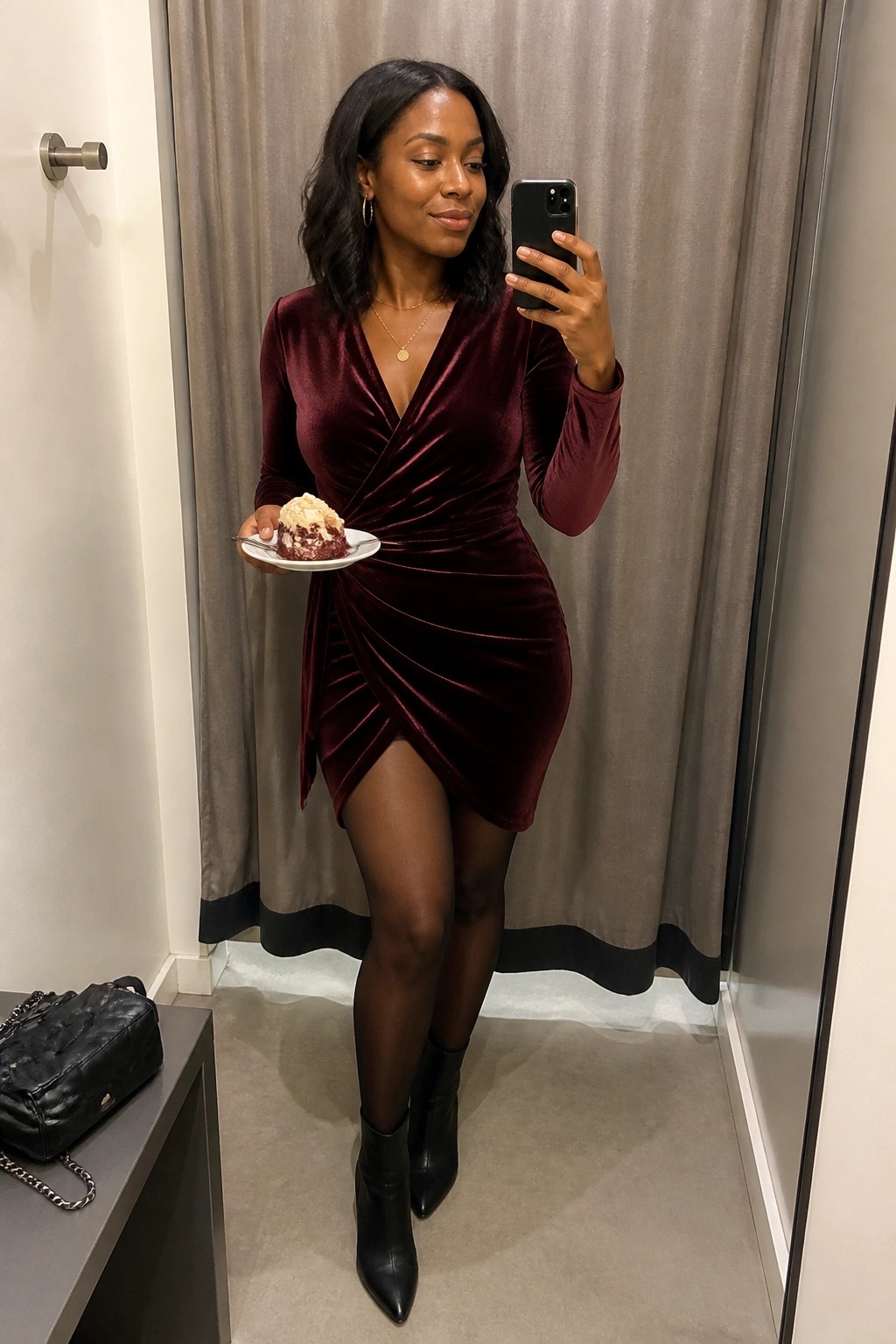 Woman in burgundy velvet dress holding cake and phone in fitting room mirror selfie.