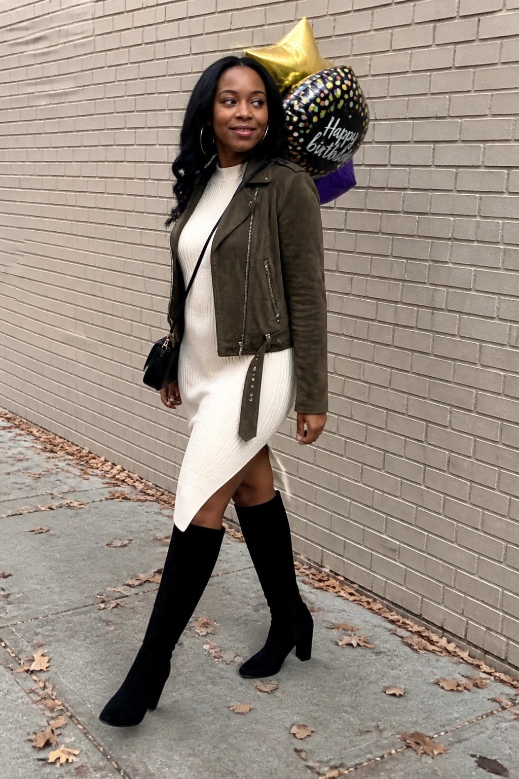 Black woman wearing a white ribbed sweater dress layered under an olive green suede moto jacket with black knee high boots and a small crossbody bag.