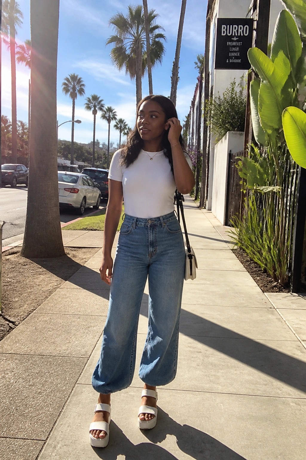 Woman wearing a white short sleeve bodysuit tucked into blue wide leg cropped jeans with white platform sandals and a small white crossbody bag.