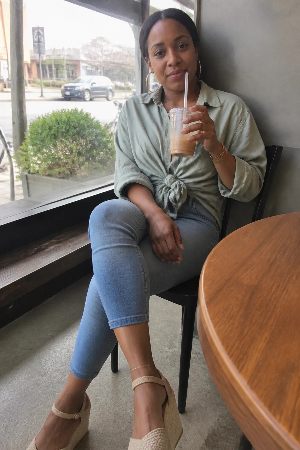 Woman in tied sage-green shirt and jeans holding iced drink in cafe