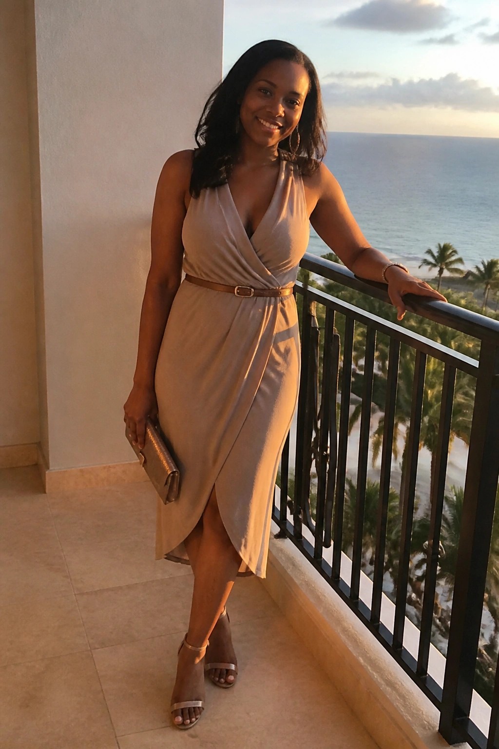 Woman in beige wrap dress smiling on ocean-view balcony at sunset