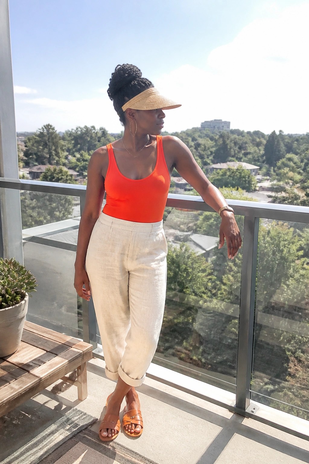 Woman in bright orange tank bodysuit, white rolled-cuff pants, brown slide sandals, straw visor, and layered gold jewelry standing on a balcony.