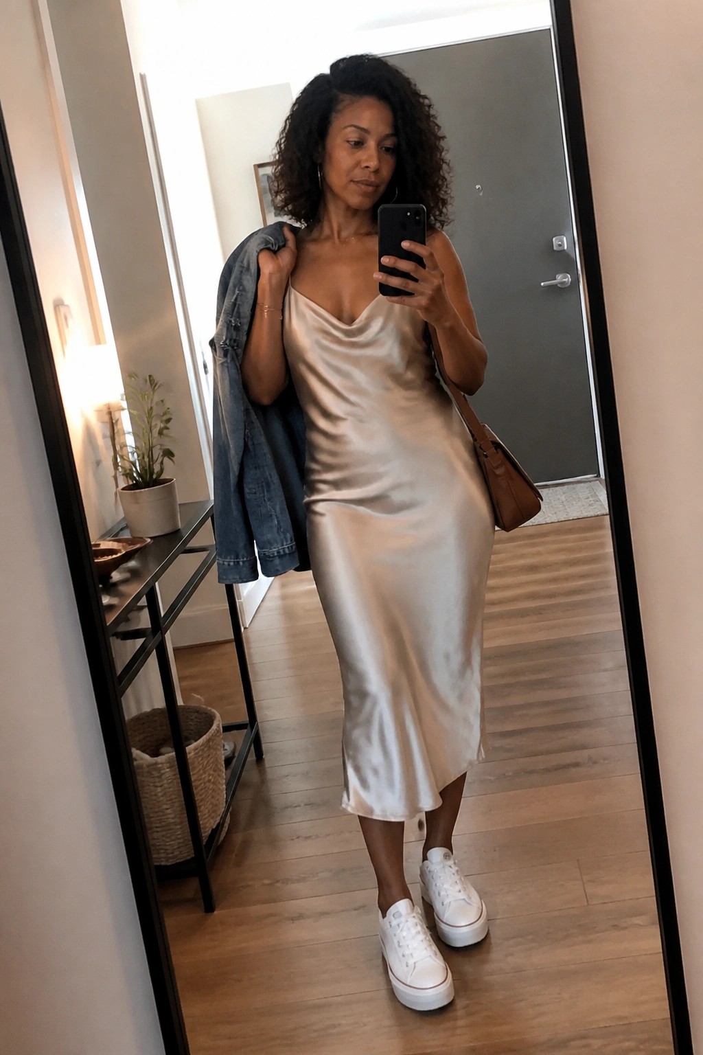 Woman wearing a champagne satin slip midi dress with white platform sneakers, holding a denim jacket and brown leather bag while taking a mirror selfie.