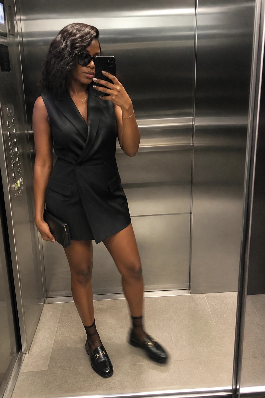 Woman wearing a black sleeveless blazer mini dress with gold buttons, paired with black patent loafers, sheer ankle socks, a black clutch, sunglasses, and delicate gold jewelry while taking a mirror selfie.