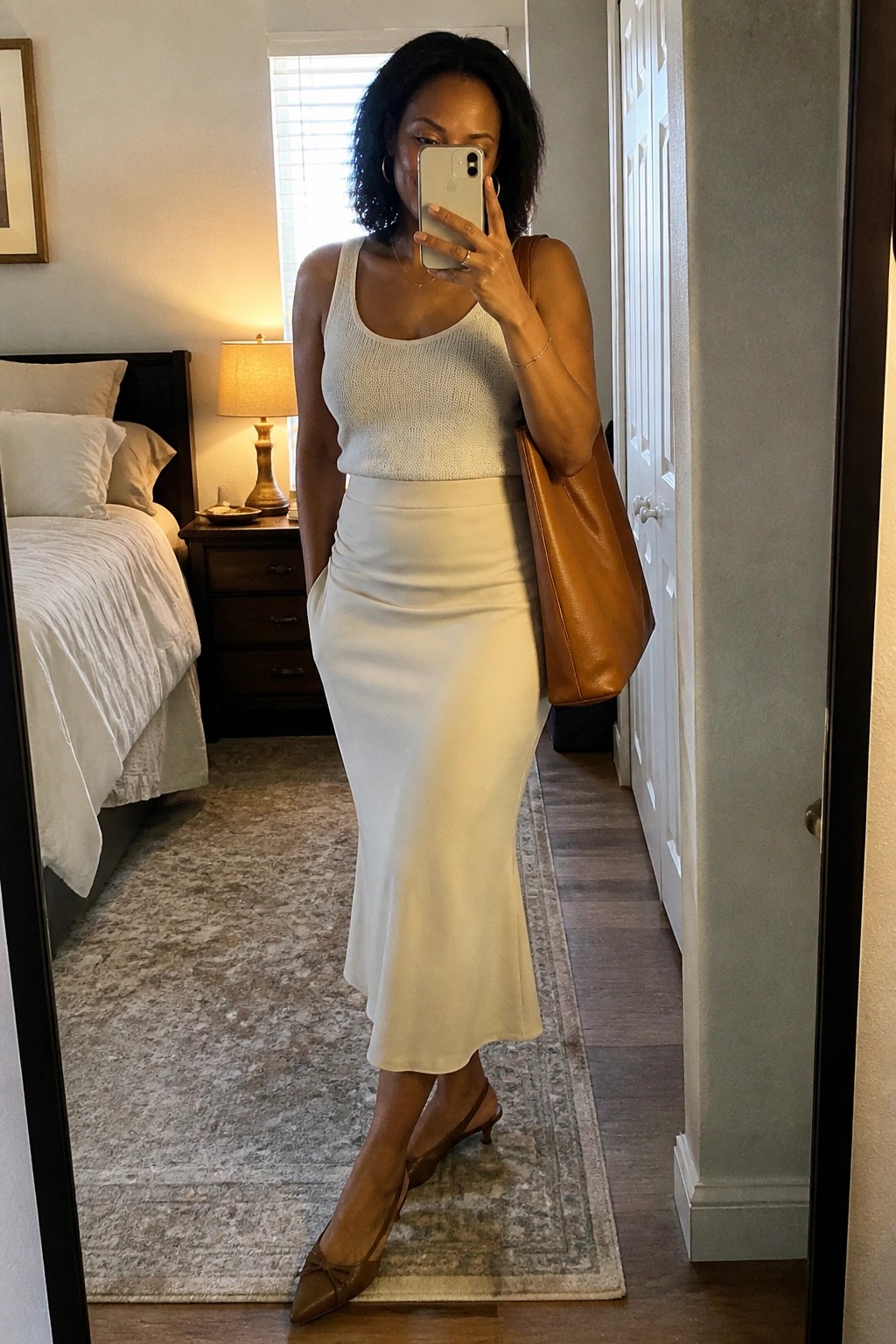 Woman in white tank top and midi skirt taking bedroom mirror selfie with brown tote