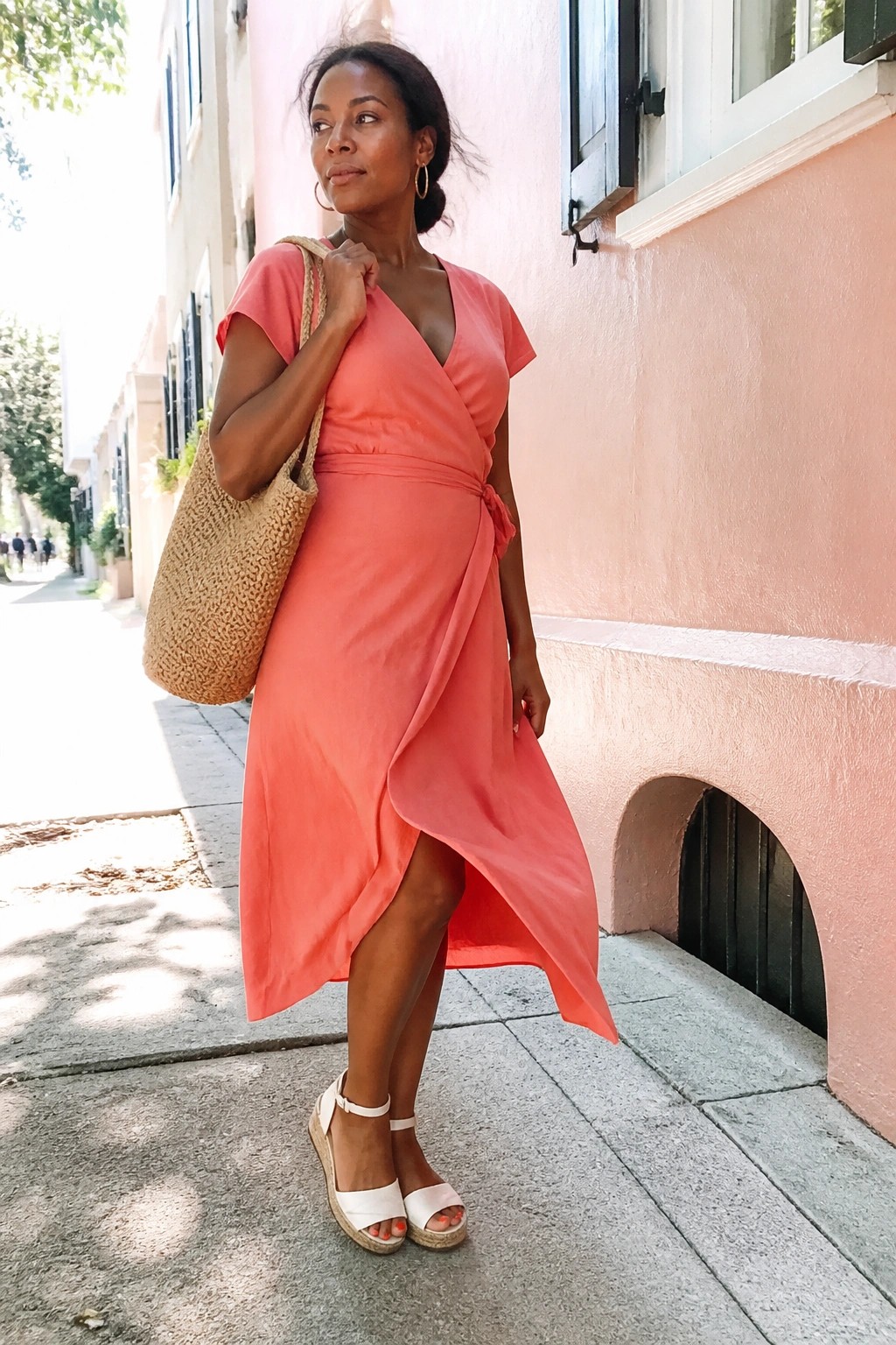 Woman in coral wrap dress holding straw tote on sunny sidewalk