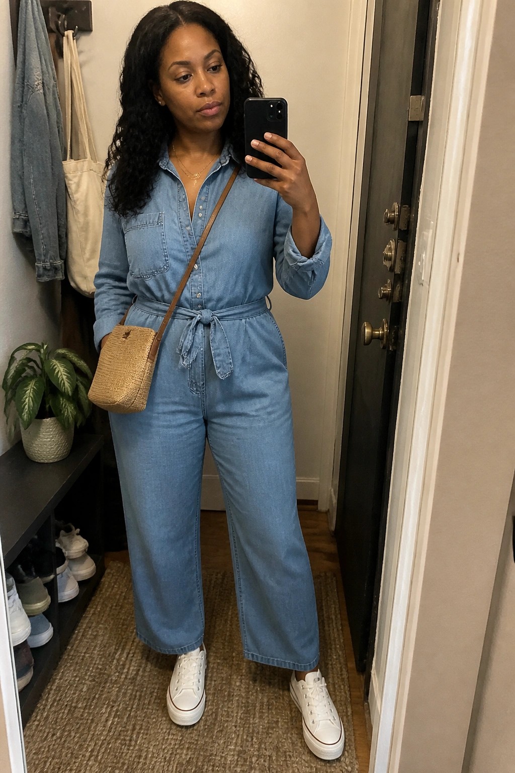 Woman in light blue denim jumpsuit taking mirror selfie with woven bag and white sneakers.