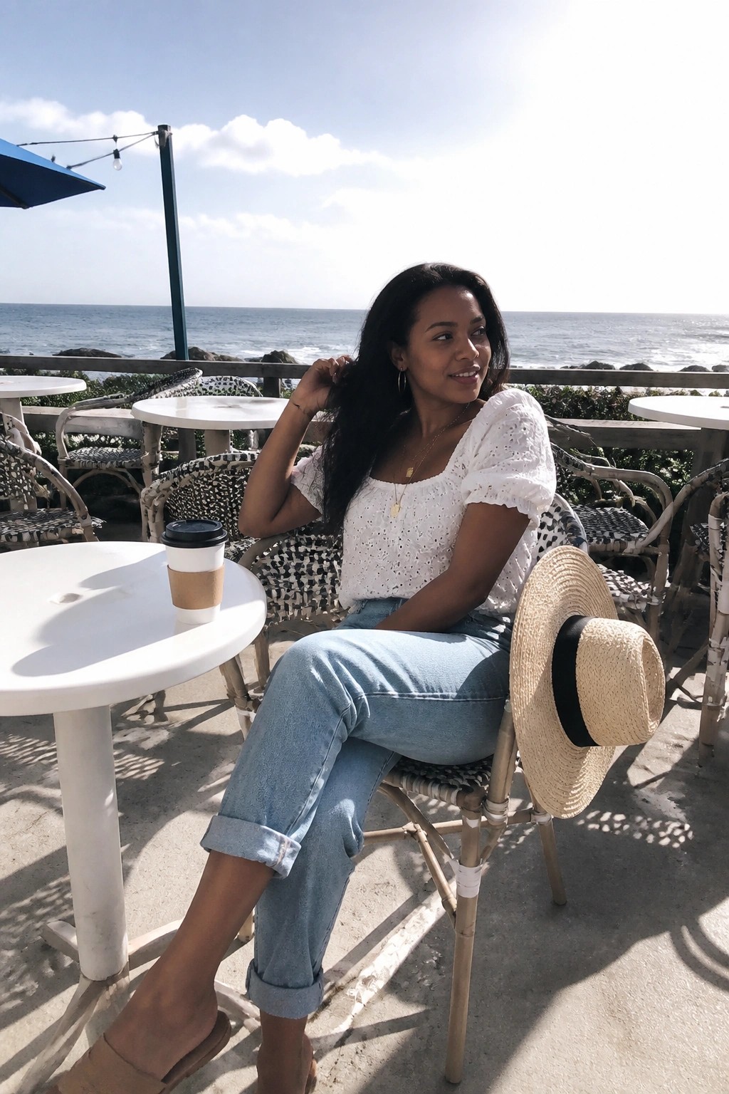 Woman in white top and jeans at ocean cafe holding straw hat beside coffee