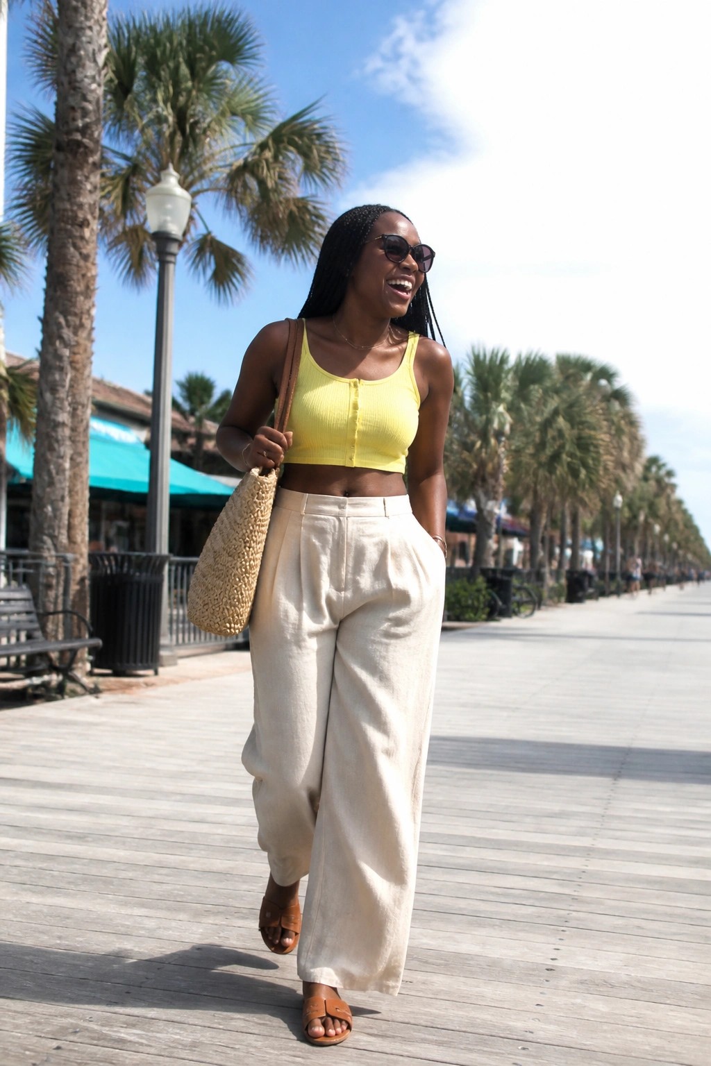 Black woman in yellow ribbed crop top and wide beige linen pants with brown leather slides and woven straw bag walking on wooden boardwalk.