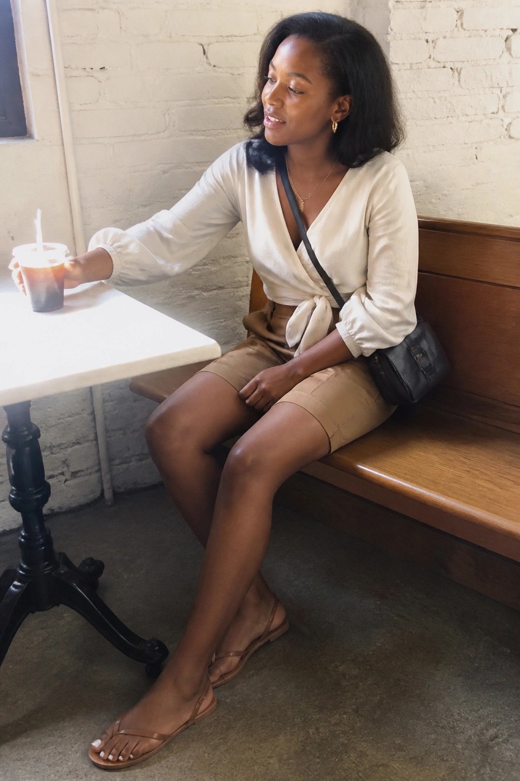 Woman in white wrap blouse and beige shorts sits on bench holding iced drink.