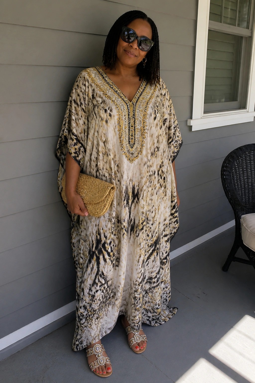 Woman in black, white, and gold patterned caftan holding gold clutch on porch.