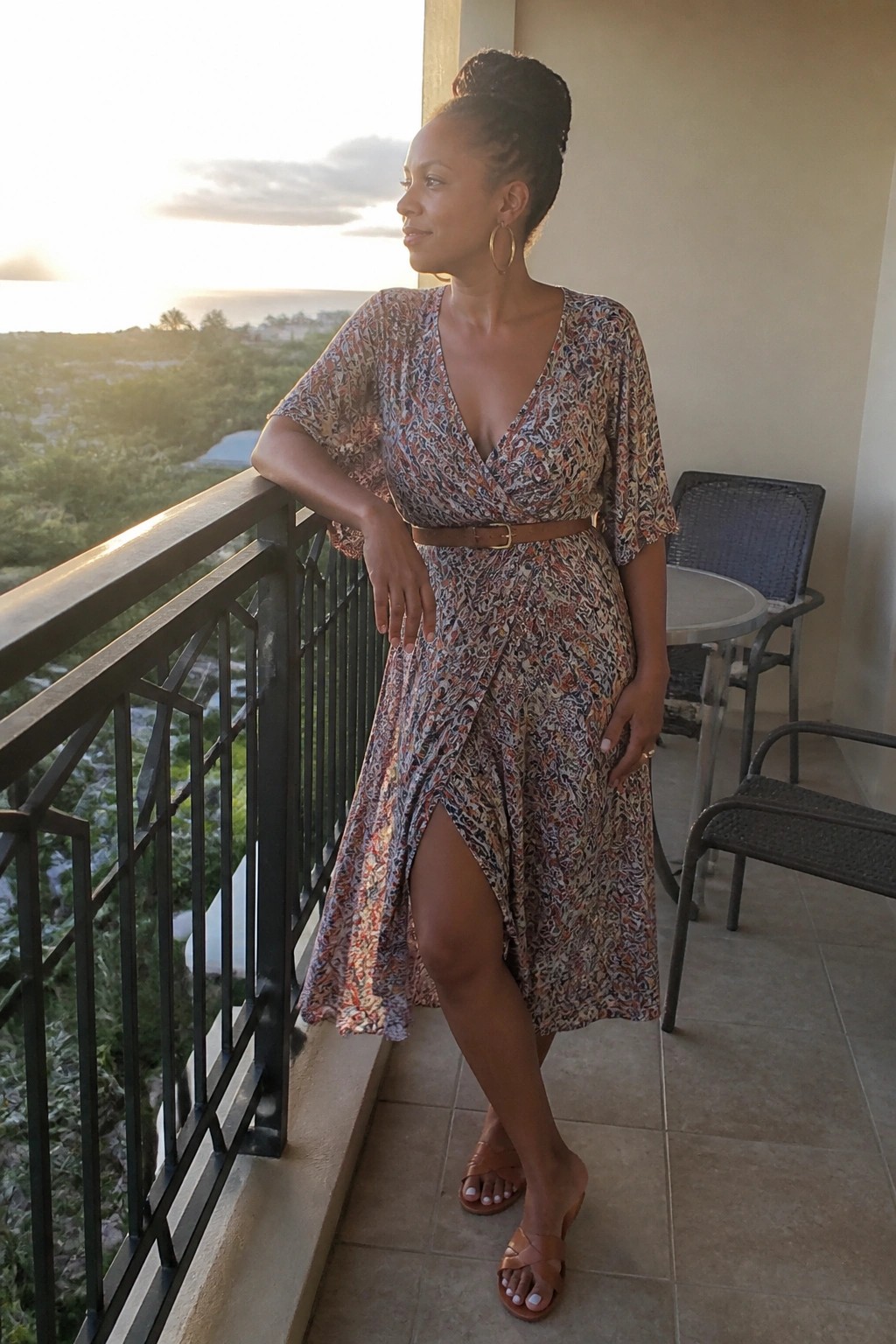 Woman wearing a multicolored printed wrap midi dress with short sleeves, brown leather belt, front slit, and brown strappy sandals on a balcony.