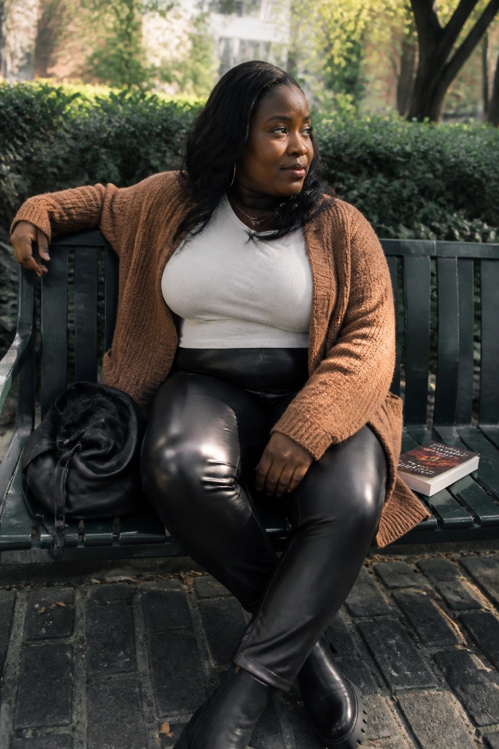 Woman in brown cardigan and black leather pants on park bench with bag and book