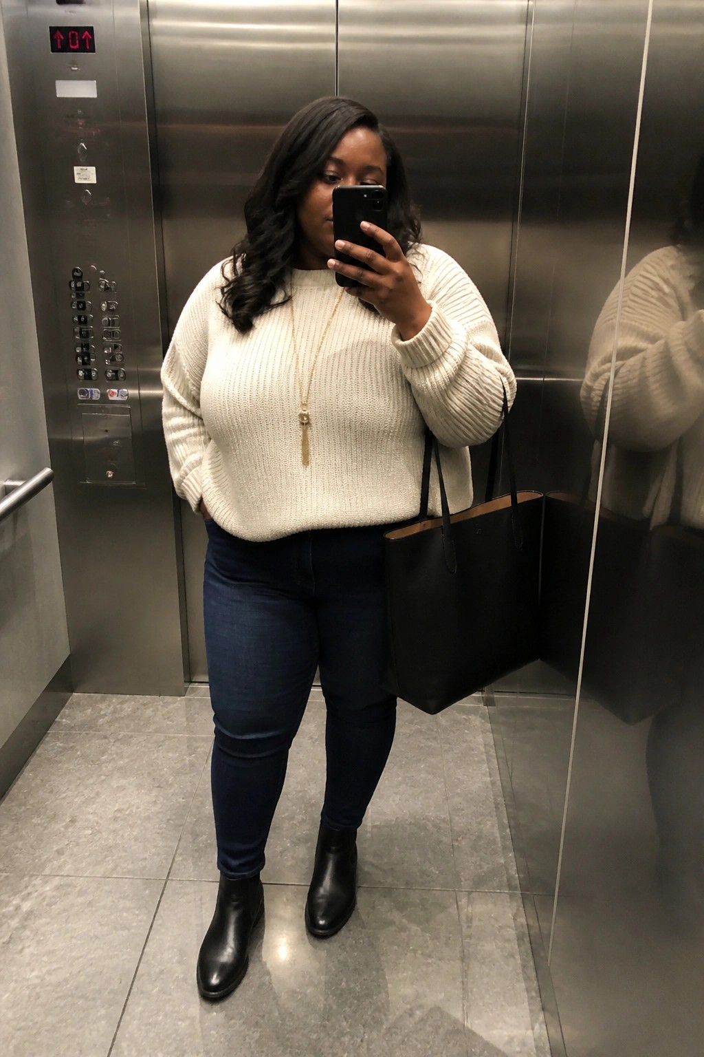 Plus size woman in an elevator mirror selfie wearing a cream chunky knit sweater, dark blue jeans, black ankle boots, and carrying a black tote bag with a long gold necklace.