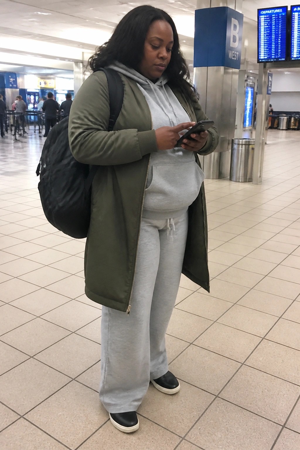 Woman in olive vest over gray sweatsuit checks phone in airport terminal