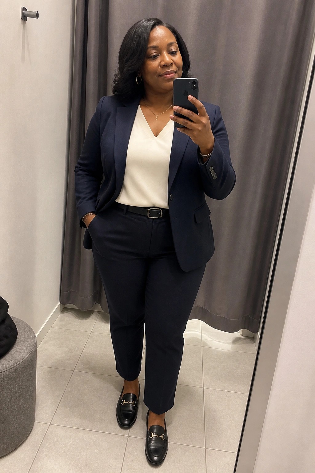 Woman in navy pantsuit taking mirror selfie in fitting room with phone