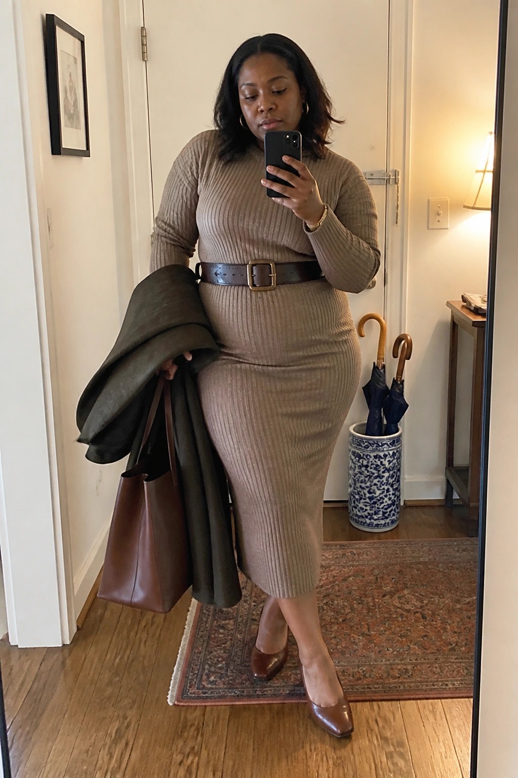 Plus size Black woman wearing a taupe ribbed knit midi dress cinched with a brown leather belt, brown pointed toe heels, and carrying a matching brown tote with a dark green jacket draped over her arm.