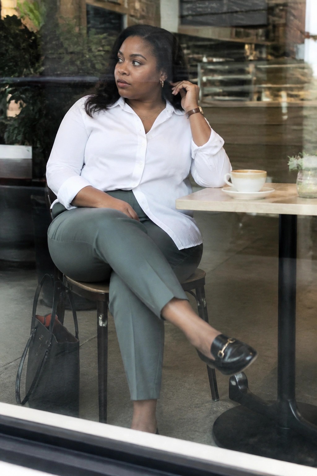 Woman in white shirt and olive pants at cafe table with coffee.