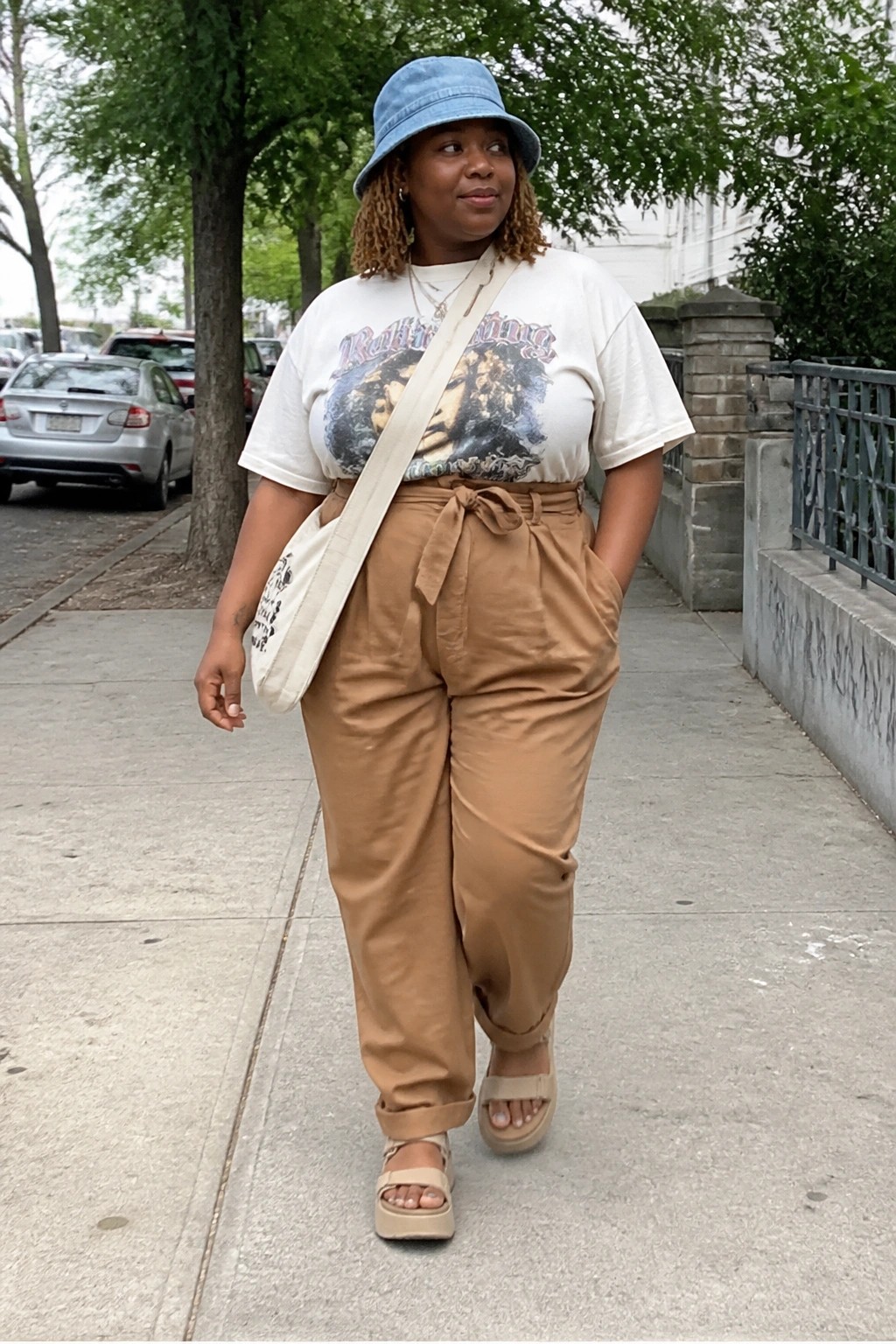 Woman in blue bucket hat, white graphic tee, brown pants, walking on sidewalk.