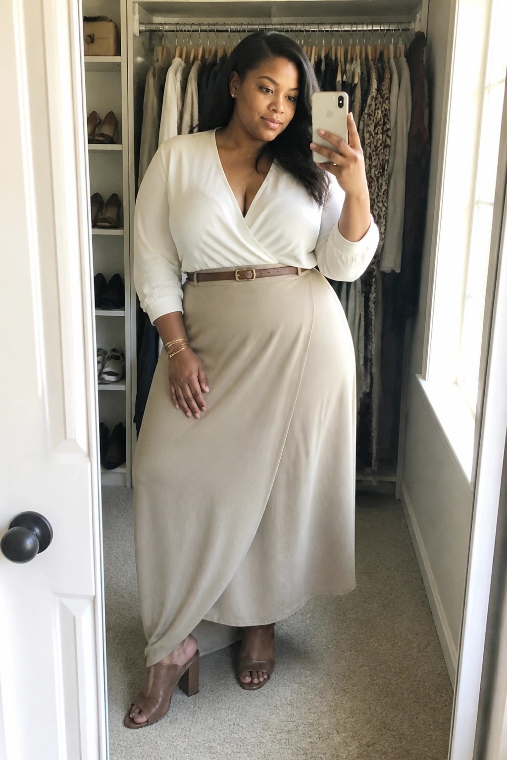 Plus size woman wearing a white wrap blouse with a beige wrap midi skirt, brown belt, and brown block heel sandals while taking a mirror selfie in a closet.