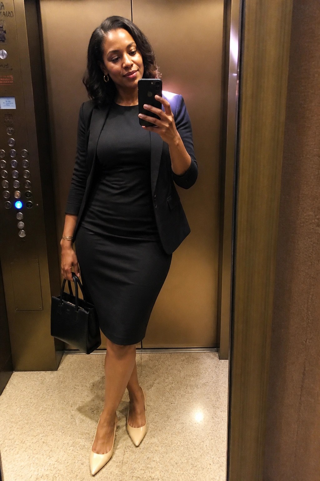 Woman in black blazer and dress taking selfie in elevator with handbag.